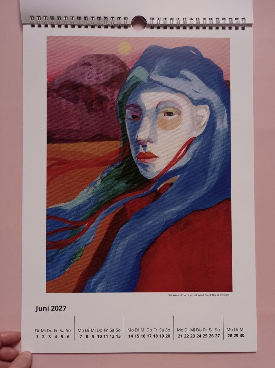 Juniblatt 2027 mit "Rückenwind": ein Frauenporträt mit wehendem blauem Haar vor einer Landschaft in Rot und Orange.